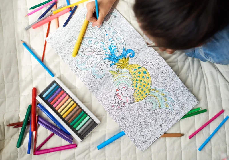 Libri da colorare per bambini. Bambino che colora un libro con disegni dettagliati, circondato da matite colorate e pastelli.